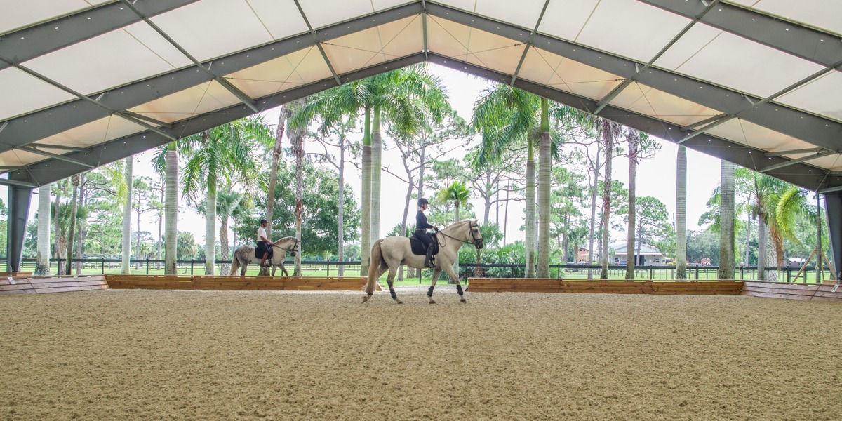 Covered equestrian arena with hybrid steel frame and fabric roof, featuring riders on horseback training in a bright, open-air environment surrounded by palm trees and scenic rural fencing.