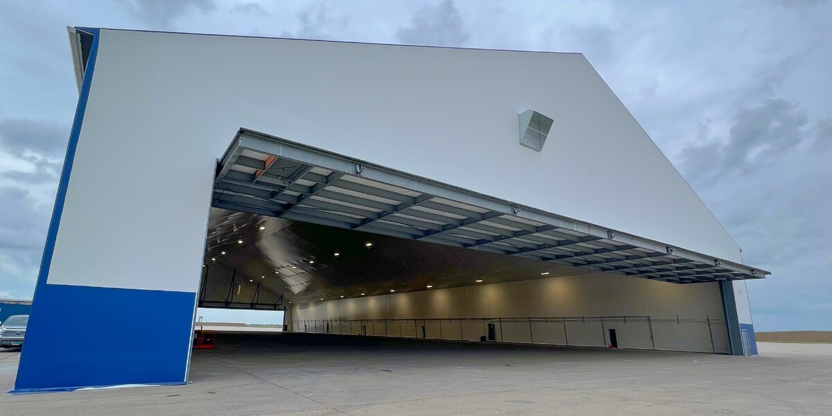 Hybrid steel aviation hangar with open fabric-clad bifold door, showcasing wide clear-span interior, integrated lighting, and modern white-and-blue exterior under a cloudy sky.
