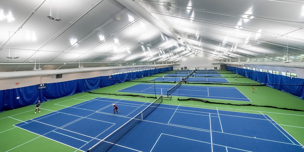 Spacious indoor tennis facility with multiple blue and green courts, bright white fabric roof, and energy-efficient lighting, featuring players in action.