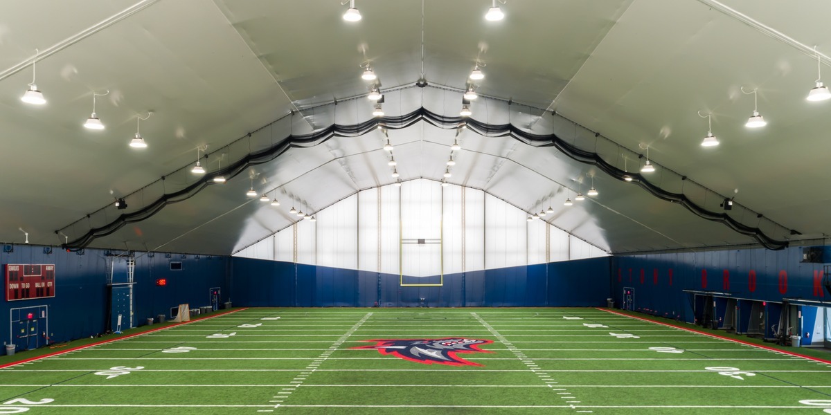 Interior view of a Stony Brook University indoor football practice facility featuring a full turf field, bright LED lighting, and a high clear-span fabric roof.