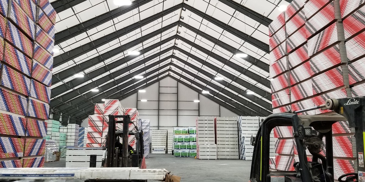 Interior of a hybrid steel fabric warehouse with high clear-span ceilings, showing stacked pallets of packaged goods and forklifts in operation under bright, evenly distributed lighting.